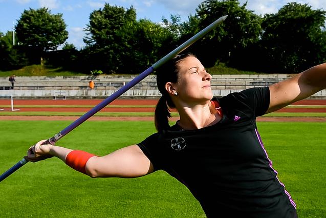 Athletin im schwarzen Trikot wirft einen Speer auf einem grünen Sportplatz bei sonnigem Wetter.