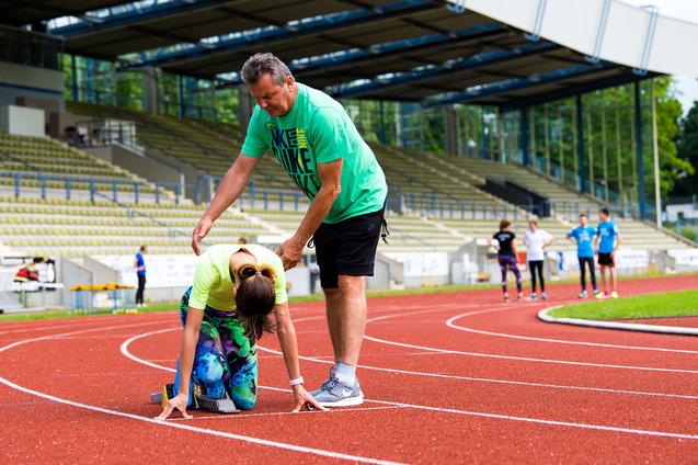 Trainer hilft Sportlerin beim Startblock auf einer Laufbahn in einem Stadion. Im Hintergrund stehen weitere Athleten.