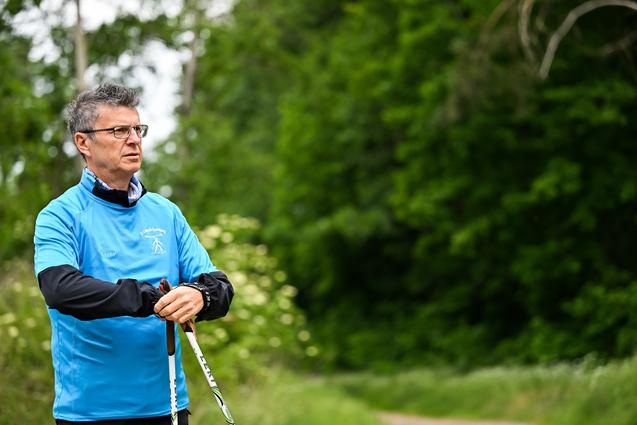 Mann in blauer Sportjacke hält Nordic Walking-Stöcke in einer grünen, bewaldeten Umgebung.