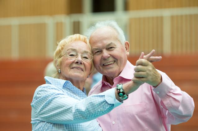 Ein älteres Paar umarmt sich lächelnd und tanzt in einer Sporthalle. Ihre Kleidung ist hell und farbenfroh.