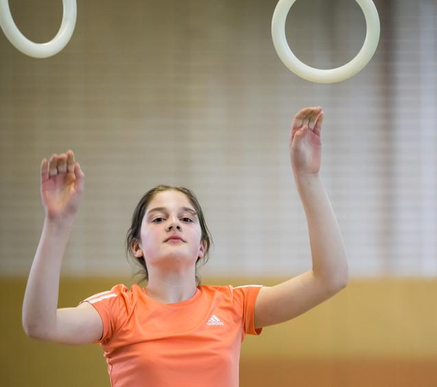 Mädchen in orangeem T-Shirt übt an Turnringen in einer Sporthalle, Hände erhoben und konzentriert.