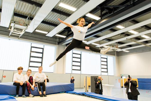 Mädchen springt mit ausgestreckten Armen über eine Trampolinfläche, während Zuschauer auf einer Bank sitzen.